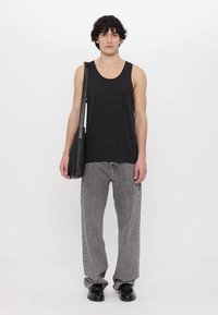 Un débardeur noir, un jean large gris et des chaussures en cuir noires. Un sac à dos noir est suspendu sur le côté. Le modèle se tient devant un fond blanc.