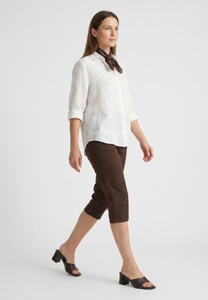 Kobieta idąca, ubrana w białą koszulę zapinaną na guziki, brązowe krótkie spodnie, czarne sandały na obcasie oraz ciemnobrązową apaszkę na szyi na jednolitym tle.