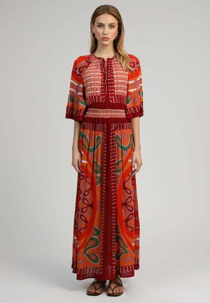 Femme debout portant une longue robe à motifs rouge, orange et vert avec des manches aux coudes et des sandales noires à brides.