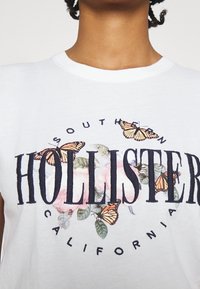 White shirt with large "Hollister" text, decorated with butterflies and floral elements, worn by a person with curly hair.