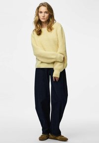 Maglione giallo chiaro lavorato a maglia con finitura testurizzata, abbinato a jeans a gamba larga blu scuro e calzature marroni. Focus sulla vestibilità comoda.