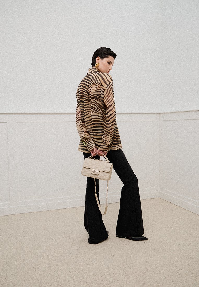 Blusa trasparente a righe marroni e nere, pantaloni neri a zampa d'elefante e una borsa crema con tracolla a catena in un ambiente minimalista.