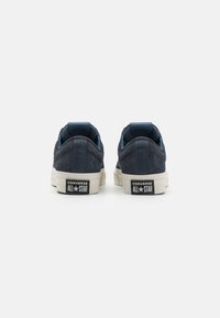Sapatilhas baixas de camurça azul-marinho com solas de borracha brancas, apresentando um design cosido e a marca "Converse All Star" no calcanhar.