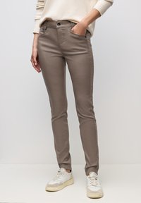 Persona che indossa pantaloni taupe aderenti e sneaker bianche, con una mano in tasca su uno sfondo chiaro uniforme.