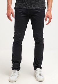 Schwarze Slim-Fit-Chinos aus Baumwolle, mit glatter Textur und subtiler Eleganz, kombiniert mit weißen Sneakers für einen Kontrast.