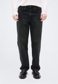 Άτομο που φοράει μαύρο τζιν με ίσιο πόδι, μαύρα δερμάτινα μοκασίνια και λευκό πουκάμισο με μακριά μανίκια, στέκεται μπροστά σε ένα απλό φωτεινό φόντο.