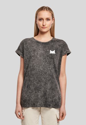 Jeune femme portant un T-shirt gris effet délavé avec un petit motif de visage de chat blanc sur la poitrine gauche et un pantalon cargo beige.