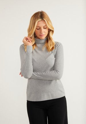 Blond kvinde iført en grå ribstrikket rullekravesweater og sorte bukser, der kigger ned med den ene hånd, der rører hendes hår.
