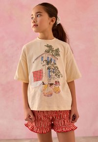 T-shirt crème avec illustration de maison et de fruits, associée à un short en vichy rouge avec ourlets à volants. Tissu texturé et coupe décontractée.