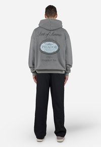 Haut à capuche gris avec un grand logo bleu au dos, affichant "Scent of Summer" et "PEGADOR". Associé à un pantalon large noir.