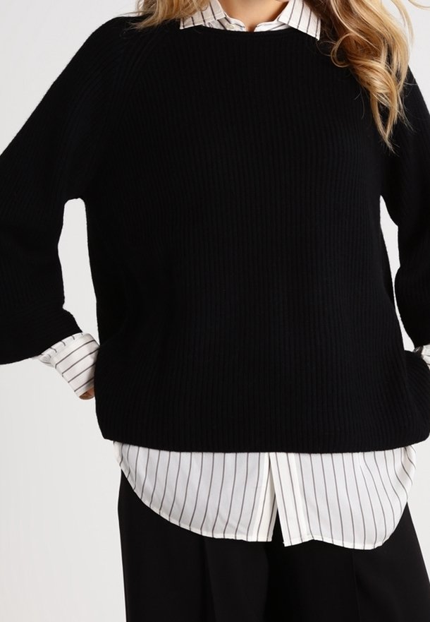 Schwarzer gerippter Strickpullover mit weiten Ärmeln, getragen über einem weißen Hemd mit dünnen schwarzen vertikalen Streifen. Das Outfit ist geschichtet und locker geschnitten.