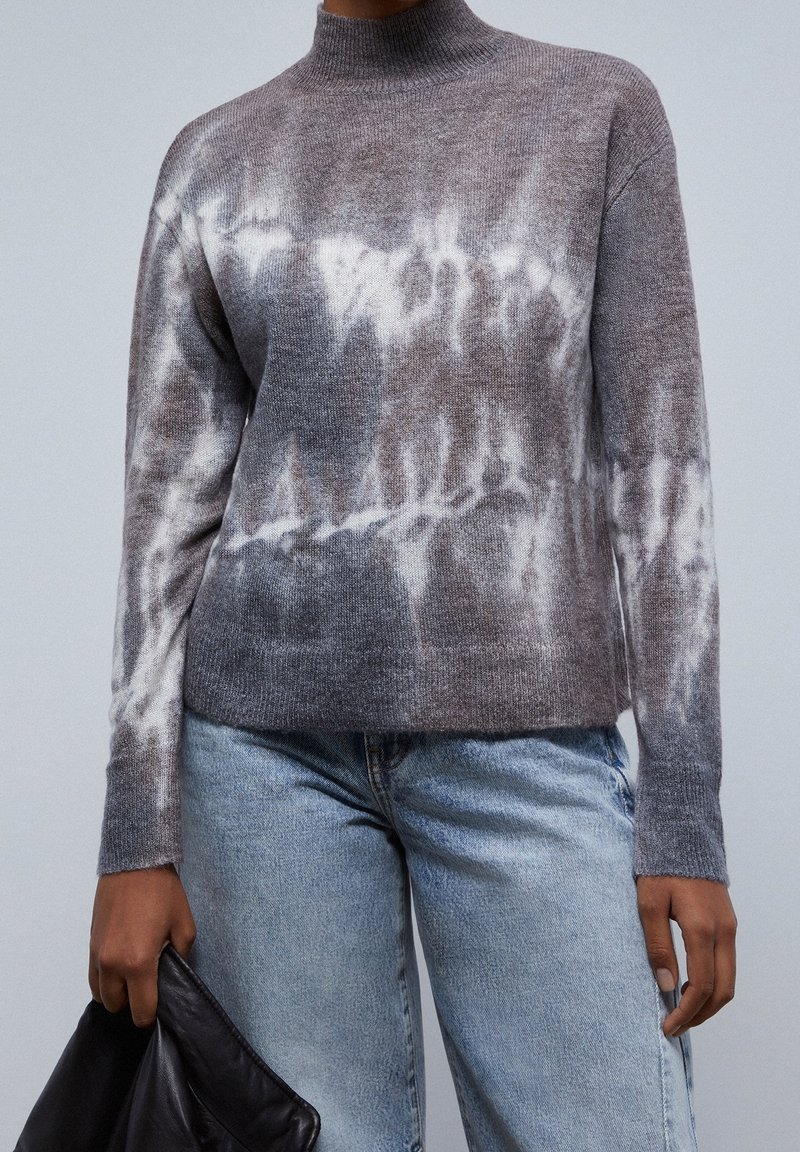 Maglione tie-dye grigio e bianco con collo alto, texture a maglia e vestibilità rilassata. Abbinato a jeans azzurri chiari e una borsa nera.