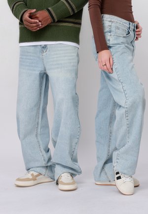 Két ember áll egymás mellett, világoskék, széles szárú farmert és laza tornacipőt viselnek; az egyikük zöld pulóvert, a másik barna felsőt.