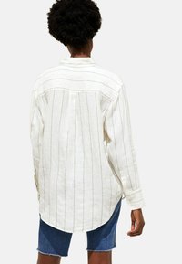 Person iført en løs hvid langærmet skjorte med tynde vertikale striber og blå denim shorts, set fra ryggen.