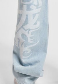 Pantalones de mezclilla azul claro con diseños gráficos blancos y patrones a lo largo de la pierna, con un ajuste holgado y detalles de costura visibles.