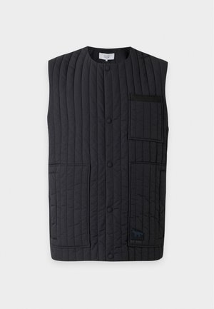 Jeune homme portant un gilet matelassé noir par-dessus une chemise grise à manches longues, un pantalon noir à jambes larges et des mocassins noir et blanc, debout devant un fond gris.