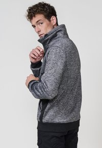 Chaqueta de forro polar texturizado en gris, con cuello alto, cierre frontal con cremallera y acentos en negro. Presenta puños y dobladillo de canalé.