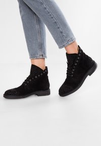 Botas negras hasta el tobillo de ante, con un exterior texturizado y remaches, cordones en la parte frontal y suela plana. Pantalones vaqueros con dobladillo recortado visible.