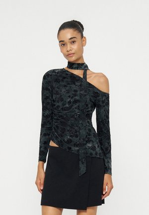 Femme portant un haut asymétrique en velours noir à motif floral avec un col ras-du-cou et une épaule dénudée, associé à une jupe noire sur fond blanc.