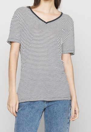 Kobieta ubrana w czarno-biały T-shirt w poziome paski z dekoltem w serek oraz niebieskie jeansy, stojąca na jednolitym tle.