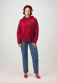 Rotes, übergroßes Hoodie, weiche Textur, mit Fronttasche, kombiniert mit hochgeschnittenen blauen Jeans. Abgerundet mit glänzenden roten High Heels.
