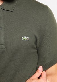 Närbild av en person som bär en mörkgrön pikétröja med en liten broderad krokodil-logotyp på bröstet.