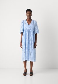 Vaaleansininen reikäommeltu mekko, jossa on syvä V-pääntie, lyhyet puffihihat ja aallotettu helma. Koristeena kukkaleikkauksia ja rento mitoitus.