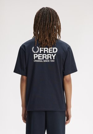 Person mit langen Dreadlocks trägt ein schwarzes T-Shirt mit dem Text "Fred Perry Original Since 1952" und einem Lorbeer-Logo auf dem Rücken.