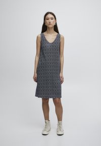 Ärmelloses V-Ausschnitt-Kleid in dunklem Navy mit einem wiederholten weißen Punktmuster, aus leichtem Stoff gefertigt, fällt knapp über das Knie. Getragen mit beigen Sneakers.