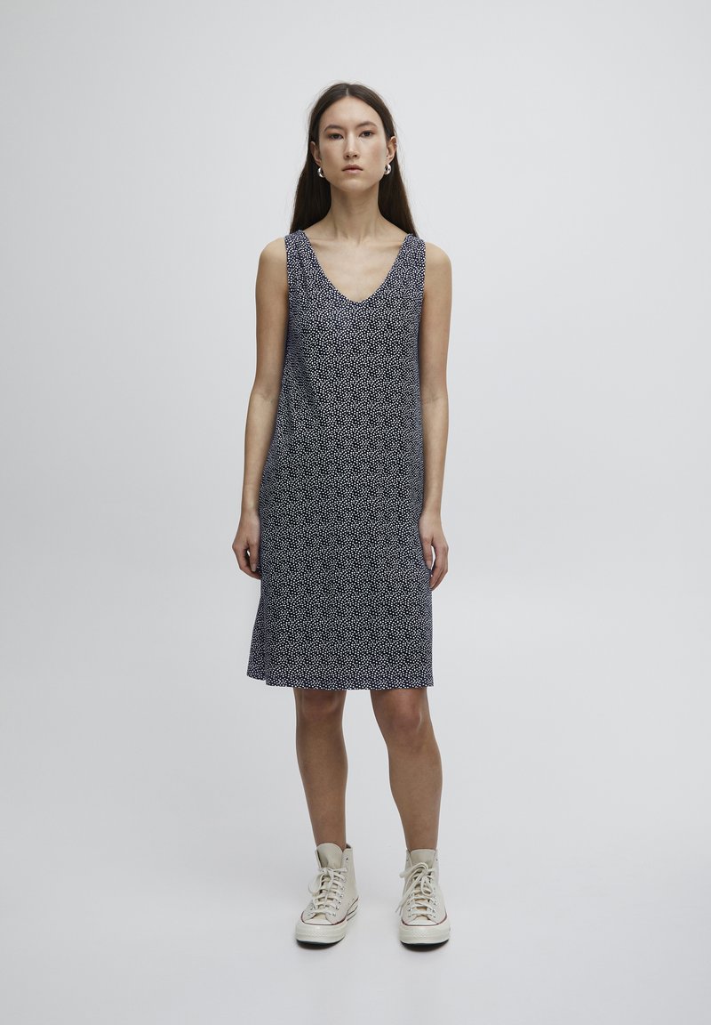 Ärmelloses V-Ausschnitt-Kleid in dunklem Navy mit einem wiederholten weißen Punktmuster, aus leichtem Stoff gefertigt, fällt knapp über das Knie. Getragen mit beigen Sneakers.