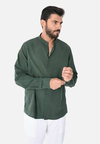 Camicia verde con colletto alla cinese, caratterizzata da una texture morbida e un design aderente. Indossata con pantaloni bianchi, maniche lunghe visibili.