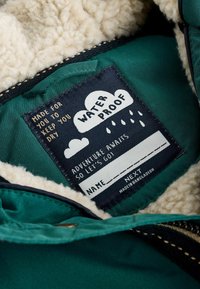 Veste imperméable verte à l'intérieur en polaire crème. L'étiquette indique "IMPERMEABLE" avec des graphismes de nuages et de gouttes de pluie.