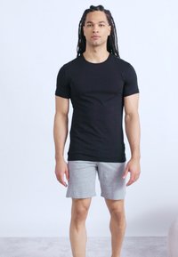 Άνδρας με μακριά πλεγμένα μαλλιά που φοράει εφαρμοστή μαύρη μπλούζα και ανοιχτό γκρι σορτς, στέκεται μπροστά σε απλό ανοιχτό φόντο.