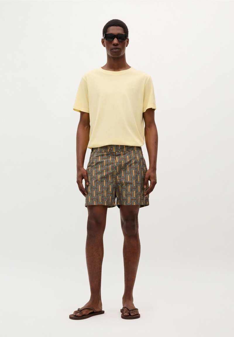Herenoutfit met een lichtgele T-shirt, gemusterde bruine shorts met rechthoekige ontwerpen en bruine slippers. Zwarte zonnebril gedragen.