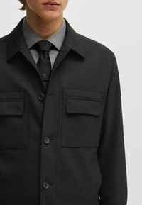 Giacca nera con bottoni, due tasche frontali, colletto e bottoni abbinati. Indossata sopra una camicia grigia e una cravatta. Texture liscia.