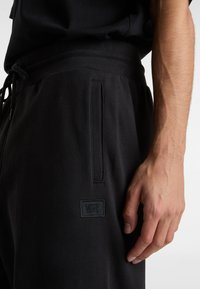 Schwarze Sweatpants mit strukturierter Oberfläche, ausgestattet mit einer Seitentasche und einem kleinen schwarzen Vans-Logopatch. Sichtbarer Kordelzugbund.
