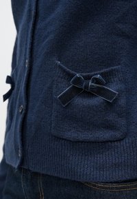 Tengerészkék kardigán texturált anyagból, elöl gombokkal és két foltzsebbel, amelyeken bársony masnik díszítik. Bordázott szegély és mandzsetták.