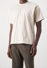 Mies, joka pukeutuu yksinkertaiseen beigeeseen lyhythihaiseen t-paitaan ja ruskeisiin viivapukuun, seisoo valkoista taustaa vasten.