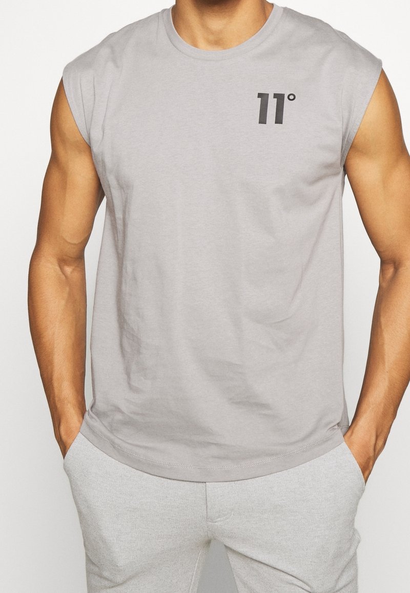 Mann in hellgrauem ärmellosem Shirt mit „11°“ Logo auf der linken Brust und passenden hellgrauen Hosen, Hände in den Taschen.