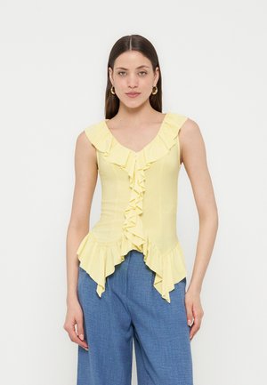 Tânără cu păr lung și castaniu, purtând cercei aurii rotunzi, un top galben fără mâneci cu volane și pantaloni albaștri largi, pe un fundal alb simplu.