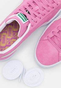 Rosa Wildleder-Sneaker mit weißen Gummisohlen und weißen Schnürsenkeln. Mit einem grünen und schwarzen Logo auf der Zunge und einer gemusterten lila Innensohle.