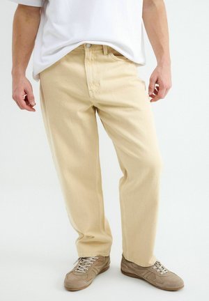 Man draagt losse beige broek, bruine vetersneakers en een wit T-shirt, staand tegen een effen achtergrond.