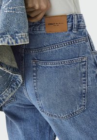 Vaqueros de denim azul con un lavado claro, que presentan una etiqueta de cuero en la cintura, dos bolsillos traseros y detalles de costura visibles.