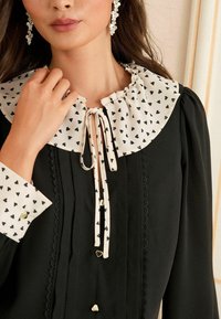 Donna che indossa un vestito nero con colletto e polsini bianchi a forma di cuore, tiene in mano la cravatta del colletto e indossa orecchini pendenti di cristallo.