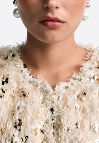 Beaded cream jacket with a textured, fringed pattern, featuring sequins, pearls, and rhinestone embellishments along the neckline.