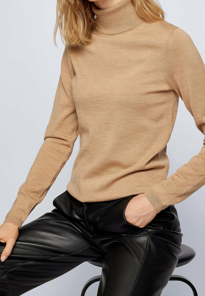 Suéter beige de cuello alto con textura acanalada y mangas largas, combinado con pantalones de cuero negros, que presentan un diseño ajustado y un cuello alto.