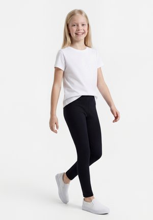Fille souriante marchant, portant un t-shirt blanc à manches courtes, des leggings noirs et des chaussures blanches sans lacets sur fond blanc.
