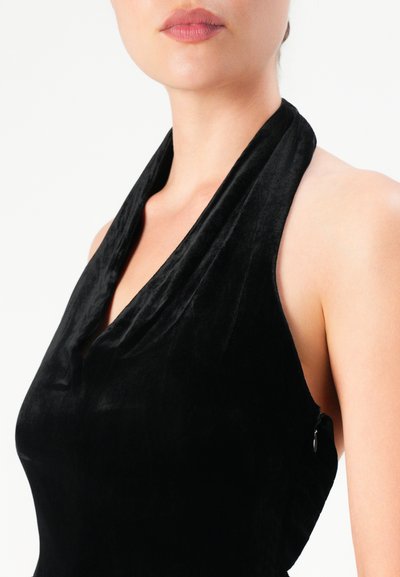 Woman wearing a black halter neck velvet dress, showing shoulders and neck against a plain white background.