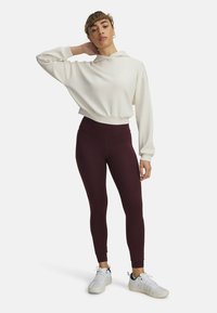 Weißer, kurz geschnittener Kapuzenpullover mit Kapuze und langen Ärmeln, kombiniert mit weinroten, hoch taillierten Leggings. Die Model trägt weiße Sneakers.