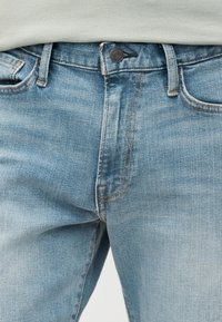 Τζιν παντελόνι από ανοιχτό μπλε τζιν στη μέση και το πάνω μέρος του μηρού με λεπτομέρειες κουμπιού και τσέπης, φορεμένο με ανοιχτόχρωμο τοπ.